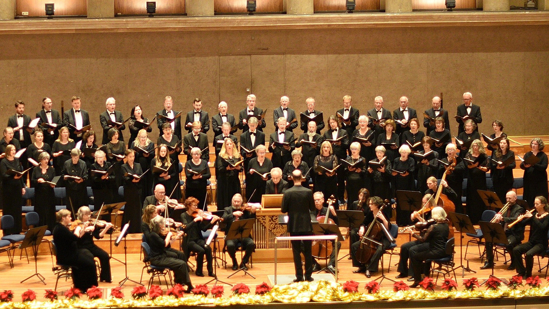 Großer Konzertchor in schwarzer Kleidung singt gemeinsam auf einer Bühne. Im Vordergrund dirigiert eine Person ein Kammerorchester mit Streichinstrumenten, während der Chor in mehreren Reihen hinter dem Ensemble aufgestellt ist.