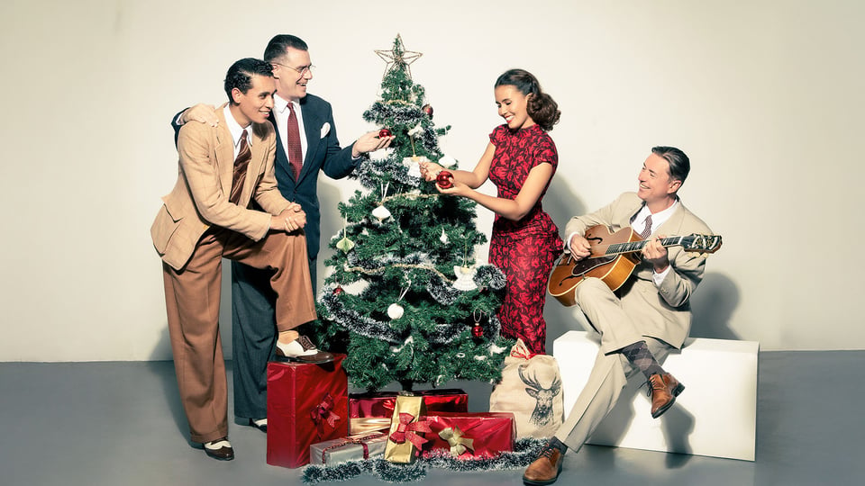 Vier Personen in eleganter Vintage-Kleidung dekorieren gemeinsam einen festlich geschmückten Weihnachtsbaum, daneben liegen Geschenke; eine Person sitzt seitlich und spielt Gitarre in heller Studio-Umgebung