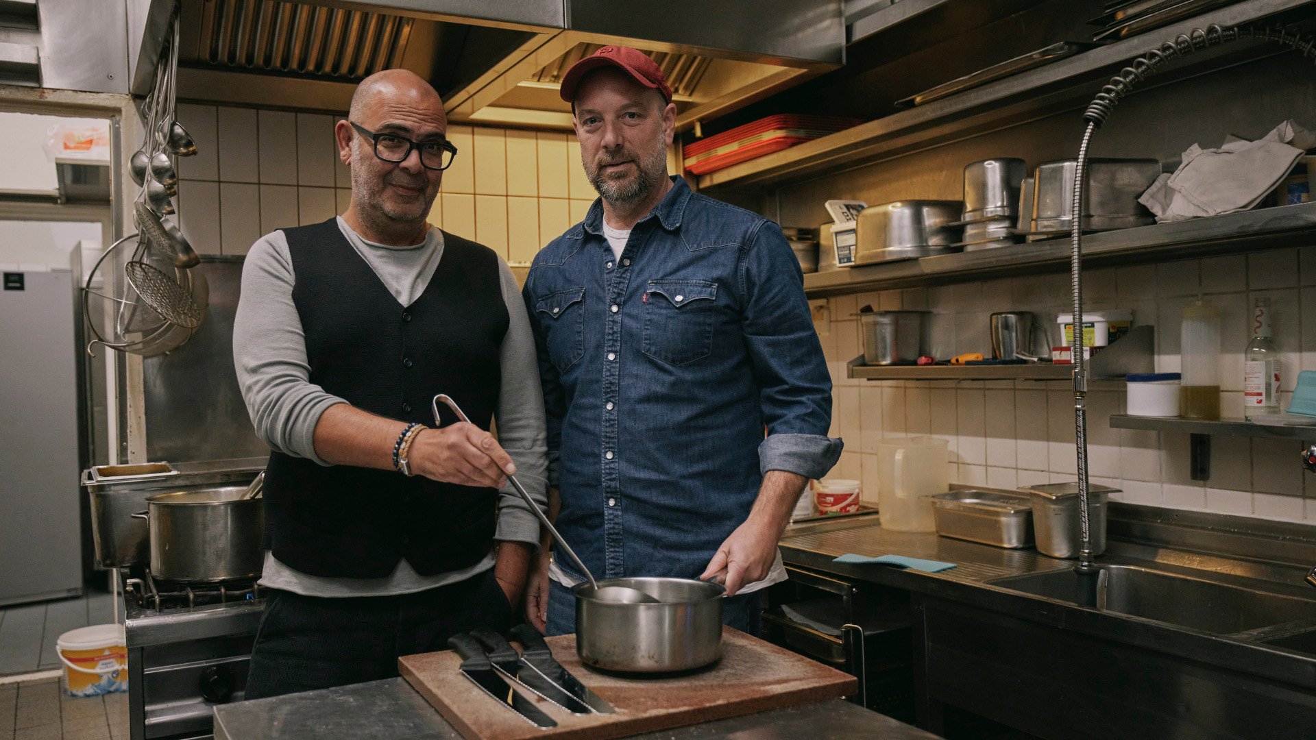 Zwei Personen stehen in einer professionellen Gastronomieküche hinter einem Arbeitstisch. Vor ihnen befindet sich ein Topf, in dem eine Person mit einer Kelle rührt. Die andere Person trägt eine rote Kappe und ein Jeanshemd. Auf der Arbeitsfläche liegen mehrere Küchenmesser. Im Hintergrund sind Edelstahlregale, Kochgeschirr, Spülbecken und weitere Küchengeräte zu sehen. Die Aufnahme vermittelt die Atmosphäre einer gemeinsamen Kochaktion in einer Restaurantküche.