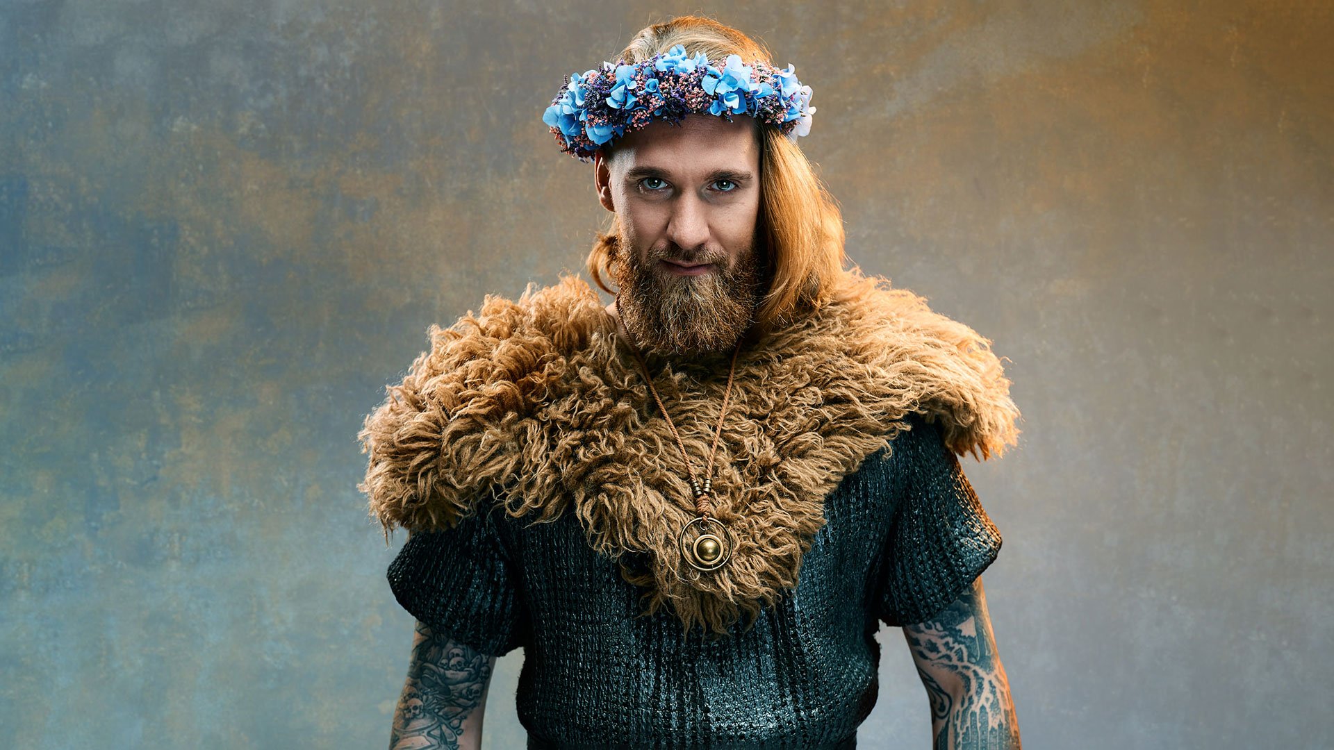 Foto für die Veranstaltung von Lennard Rosar – Schildmaid mit dem Künstler, der als Wikinger verkleidet ist und Blumen auf dem Kopf trägt, vor einem Hintergrund mit marmorierter blau‑orangefarbener Textur