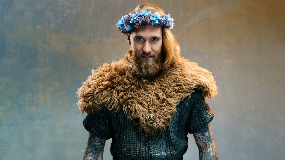 Foto für die Veranstaltung von Lennard Rosar – Schildmaid mit dem Künstler, der als Wikinger verkleidet ist und Blumen auf dem Kopf trägt, vor einem Hintergrund mit marmorierter blau‑orangefarbener Textur