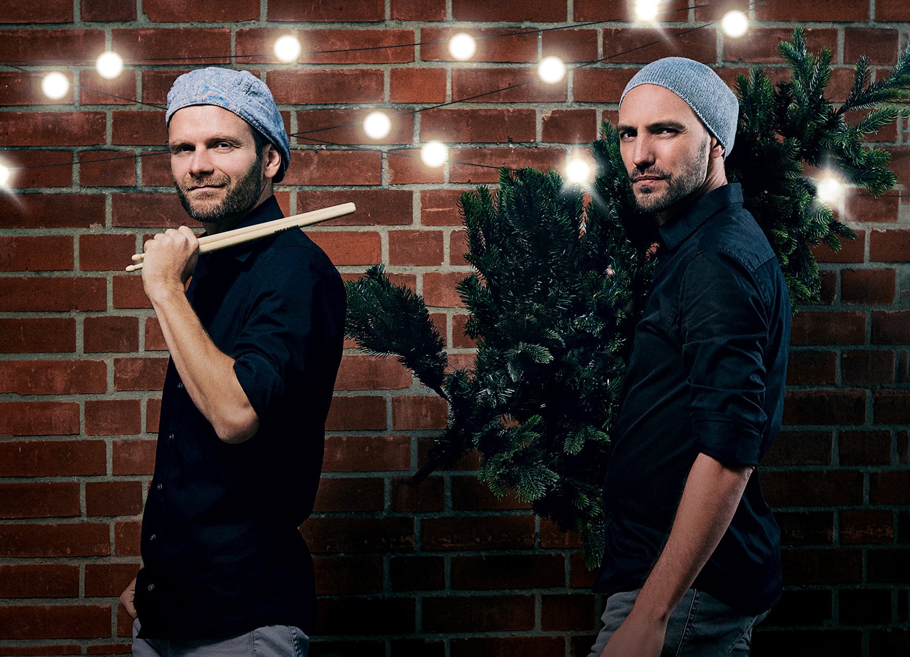 Zwei Männer in schwarzen Hemden mit blauen Kappen, einer mit Drumsticks in der Hand vor Ziegelwand