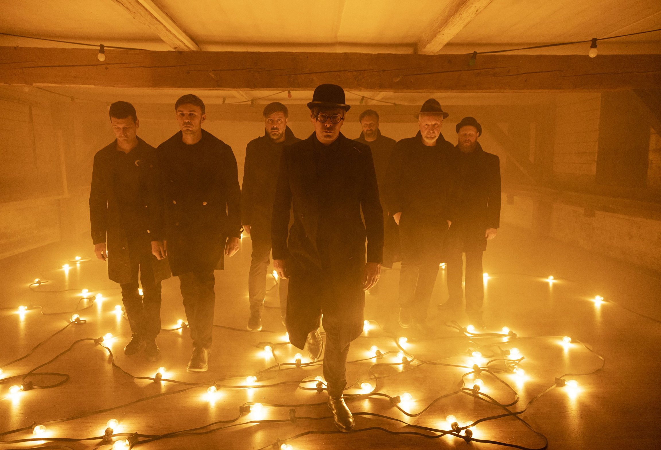 Bandfoto Kafka Band mit sechs Personen in dunkler Kleidung, die durch einen neblig beleuchteten Raum gehen, umgeben von auf dem Boden verteilten Glühbirnen und warmem orangefarbenem Licht