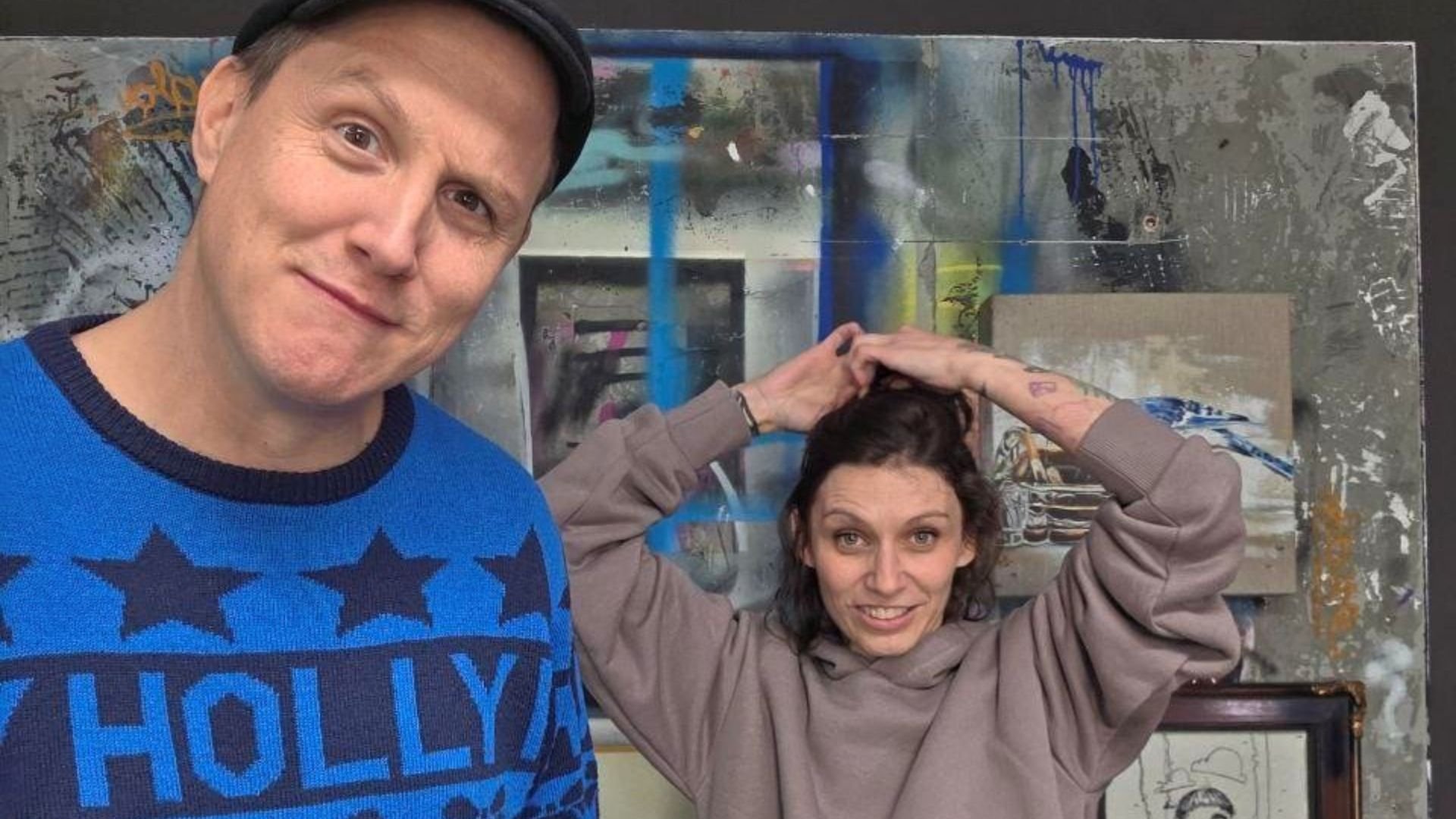 Zwei Personen vor einer bemalten Wand mit Kunstwerken; links eine Person mit Mütze und blauem Pullover, rechts eine Person mit grauem Kapuzenpulli, die sich die Haare bindet