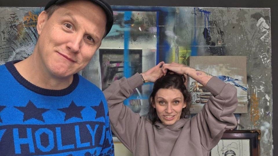 Zwei Personen vor einer bemalten Wand mit Kunstwerken; links eine Person mit Mütze und blauem Pullover, rechts eine Person mit grauem Kapuzenpulli, die sich die Haare bindet