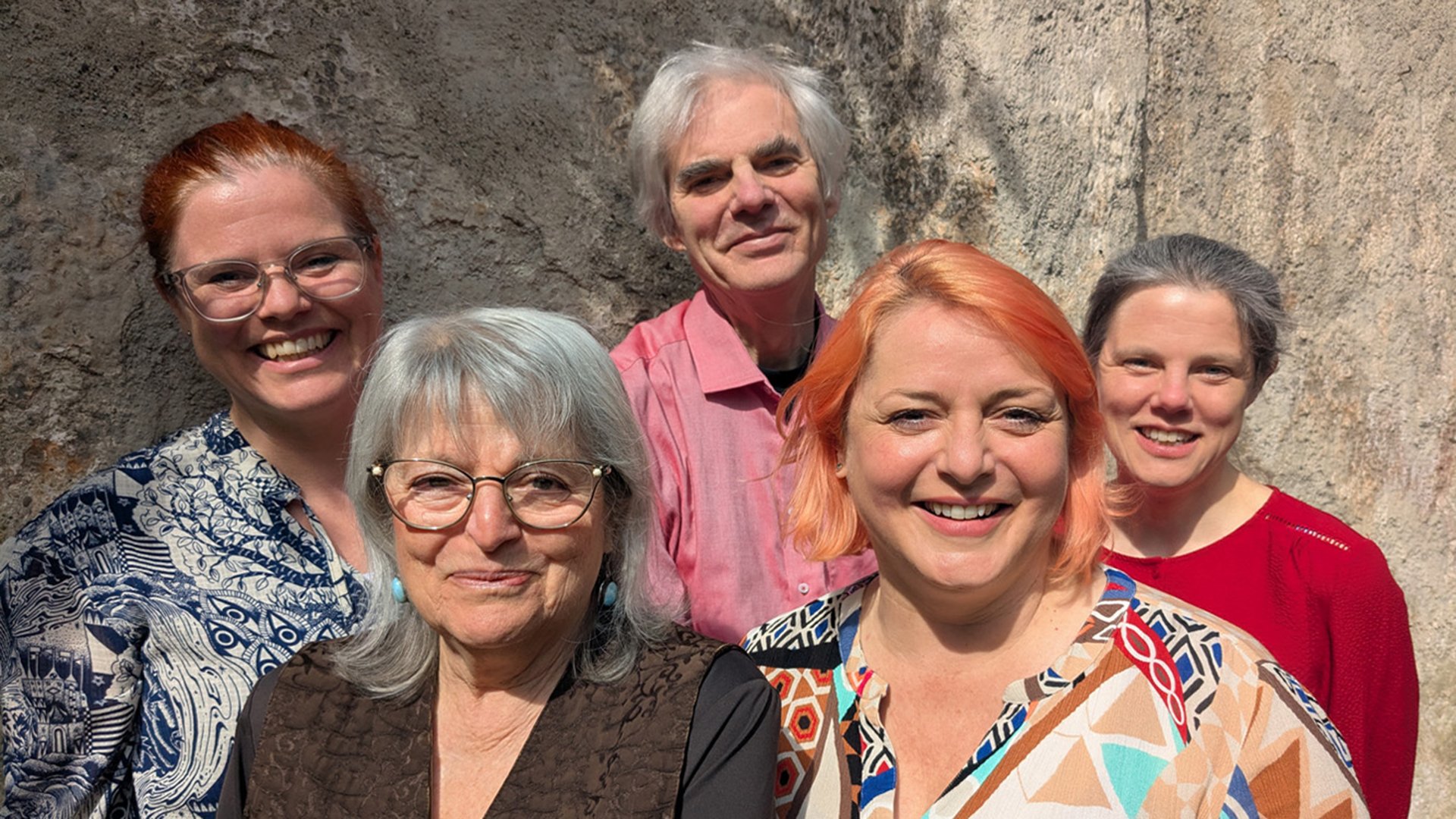 Fünf Personen in farbiger Freizeitkleidung lachend vor einer hellbraunen Steinwand