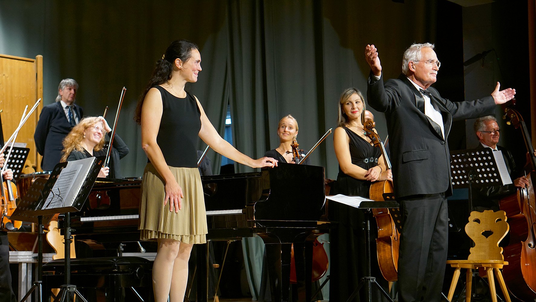 Musikerinnen und Musiker stehen mit Dirigent und Flügel auf einer Bühne und nehmen den Applaus eines Konzerts entgegen.