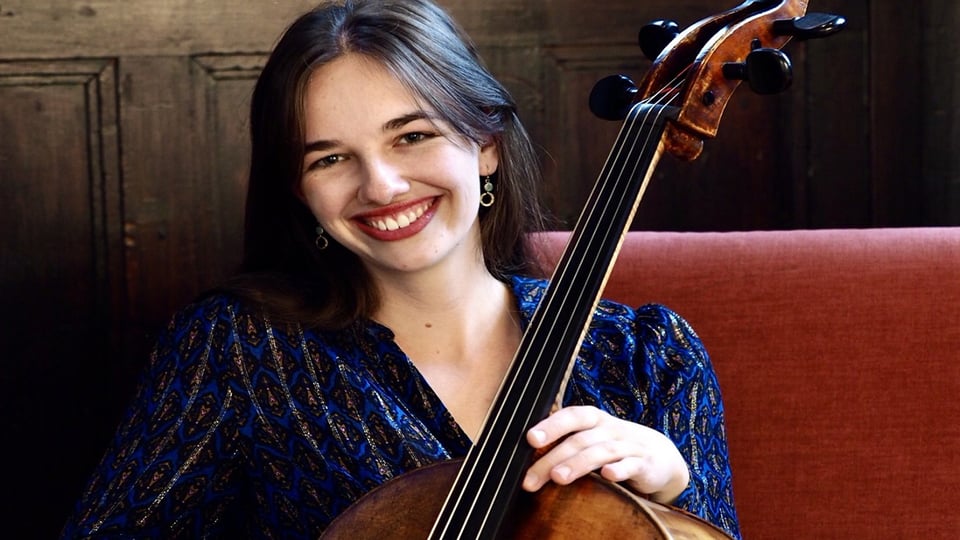 Porträt einer Cellistin mit ihrem Instrument vor dunkler Holzwand; die Person hält das Cello und sitzt vor gepolsterter Rückenlehne, warm ausgeleuchtet.