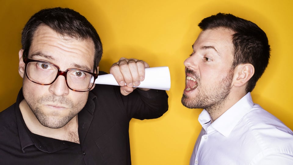 Flo und Wisch vor einem gelben Hintergrund; einer der Künstler hält ein zusammengerolltes Papier ans Ohr, während der andere daneben laut ruft