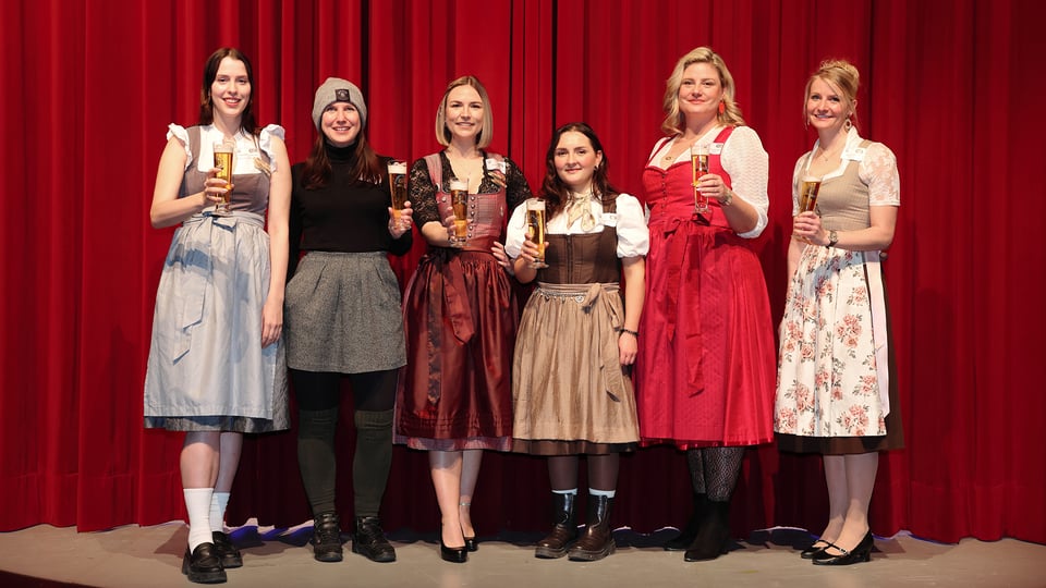 Sechs Damen in bayerischer Tracht stehen vor rotem Vorhang und halten jeweils ein Bierglas in der Hand