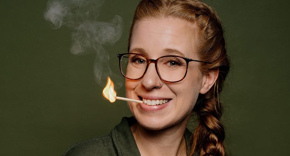 Dame mit Zopf und Brille und brennendem Steichholz im Mund vor grünem Hintergrund