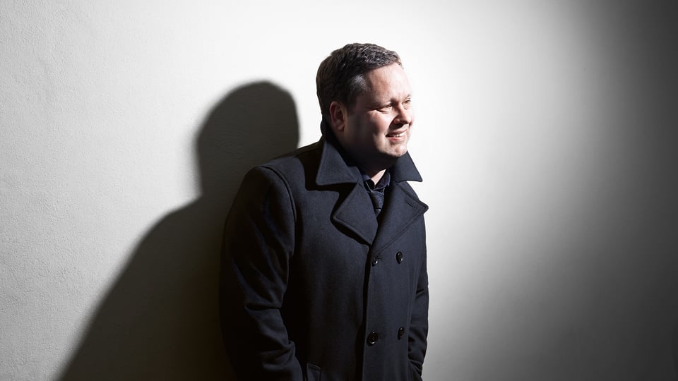 Paul Potts, britischer Tenor, in einem Porträt mit dunklem Mantel vor heller Wand, seitlich beleuchtet, mit deutlichem Schatten – Pressebild zur Tour „Unforgettable Moments“