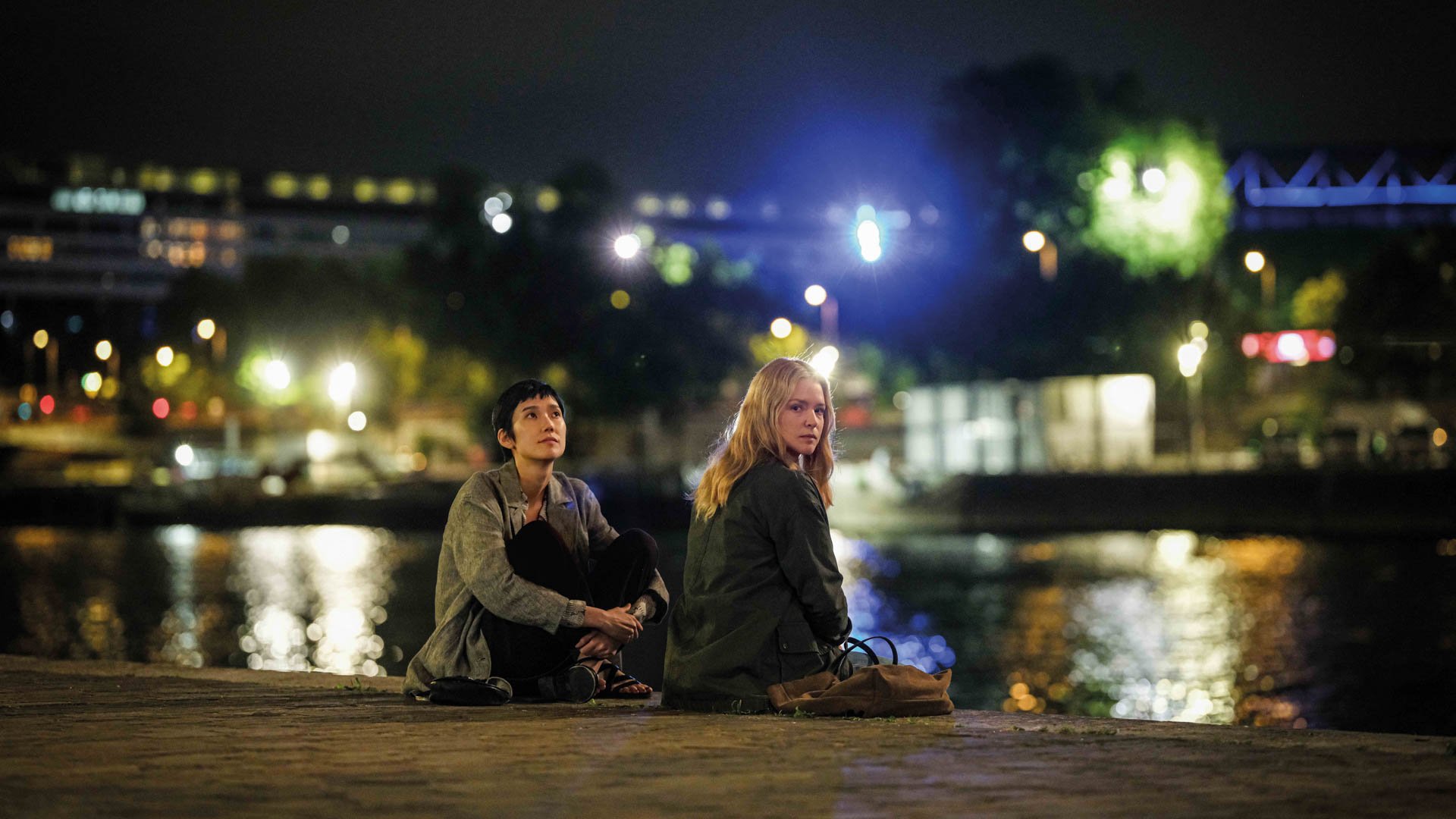 Zwei Personen sitzen nachts am Ufer eines Gewässers, im Hintergrund Lichter einer Stadt, die sich im Wasser spiegeln.