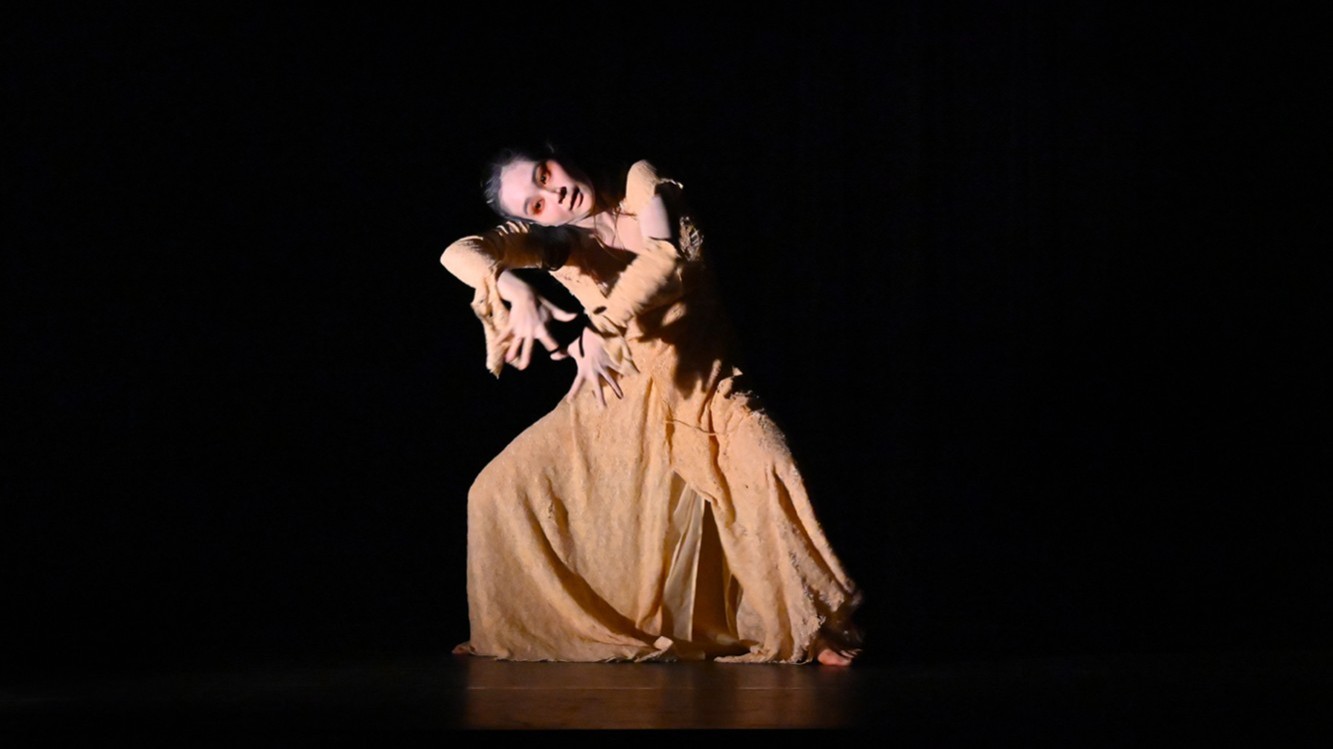 Eine Dame in einem langen, hellen Gewand tanzt allein auf einer dunklen Bühne, dramatisch beleuchtet, mit ausdrucksvoller Körperhaltung.