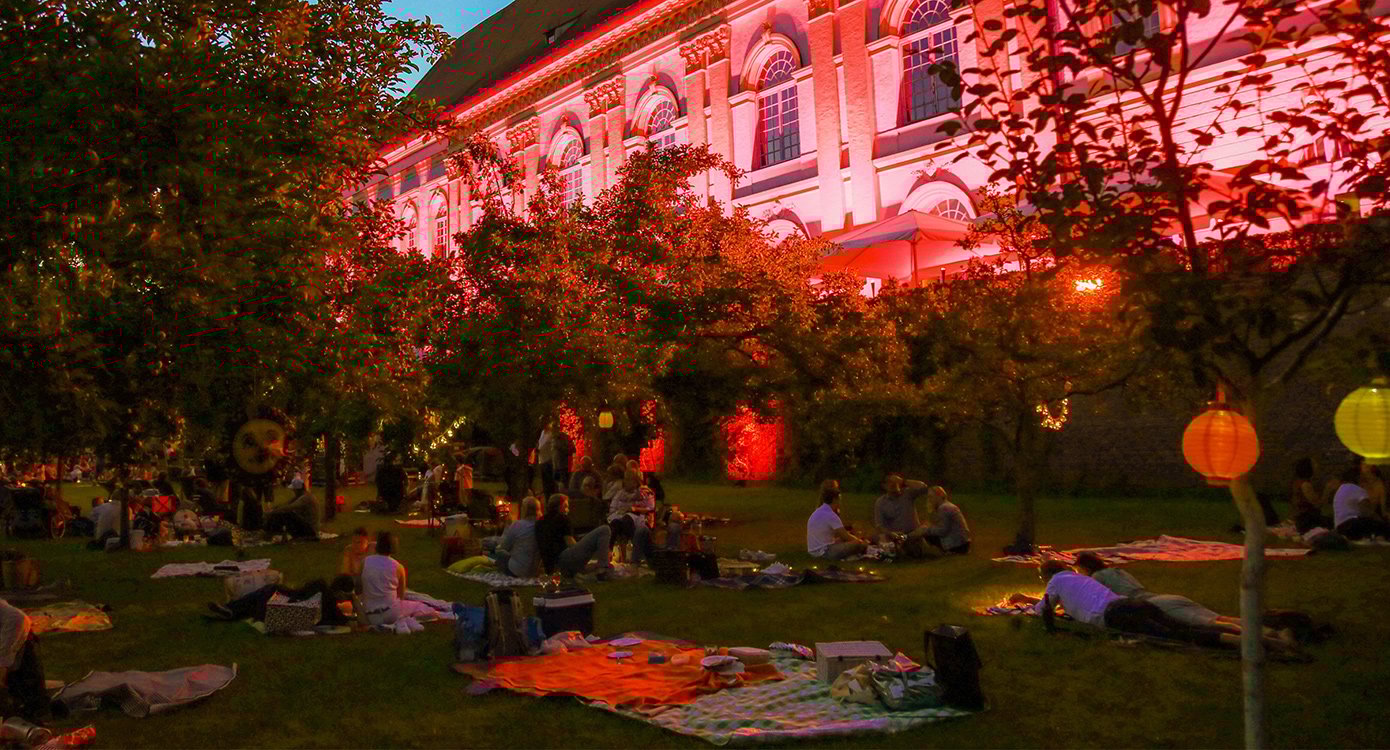Gruppe von Menschen, die auf einer Wiese Picknick machen, im Hintergrund ein historisches Gebäude