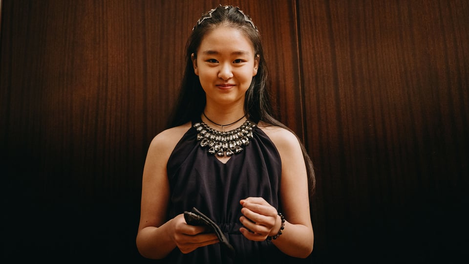Die Pianistin Tianyao Lyu in schwarzer festlicher Kleidung mit dominanter Halskette lächelnd vor braunem Hintergrund