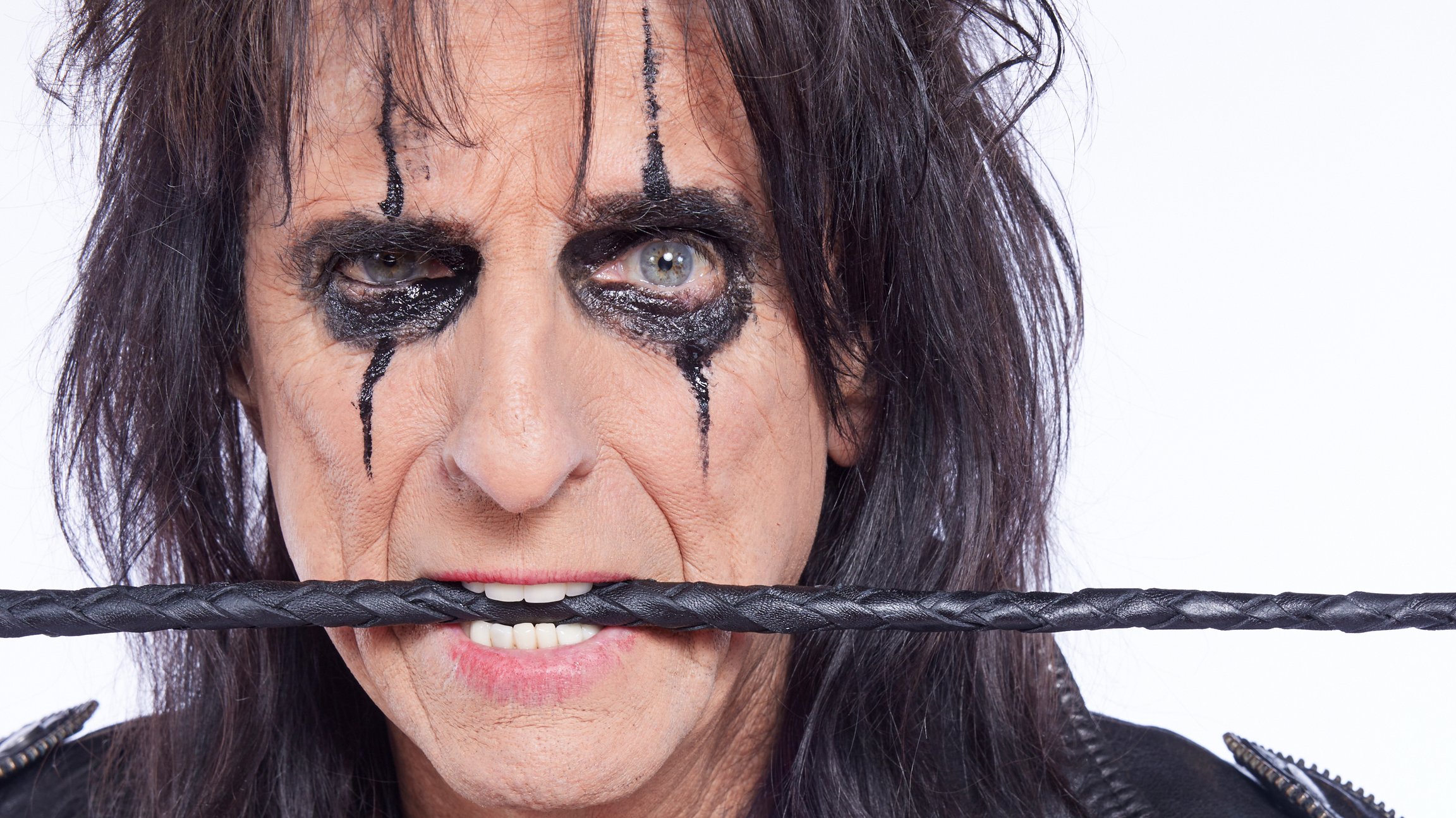 Porträtfoto von Alice Cooper in schwarzer Lederjacke vor hellem Hintergrund, mit dunklem Augen-Make-up und einem schwarzen Kabel oder Seil zwischen den Zähnen in Nahaufnahme.