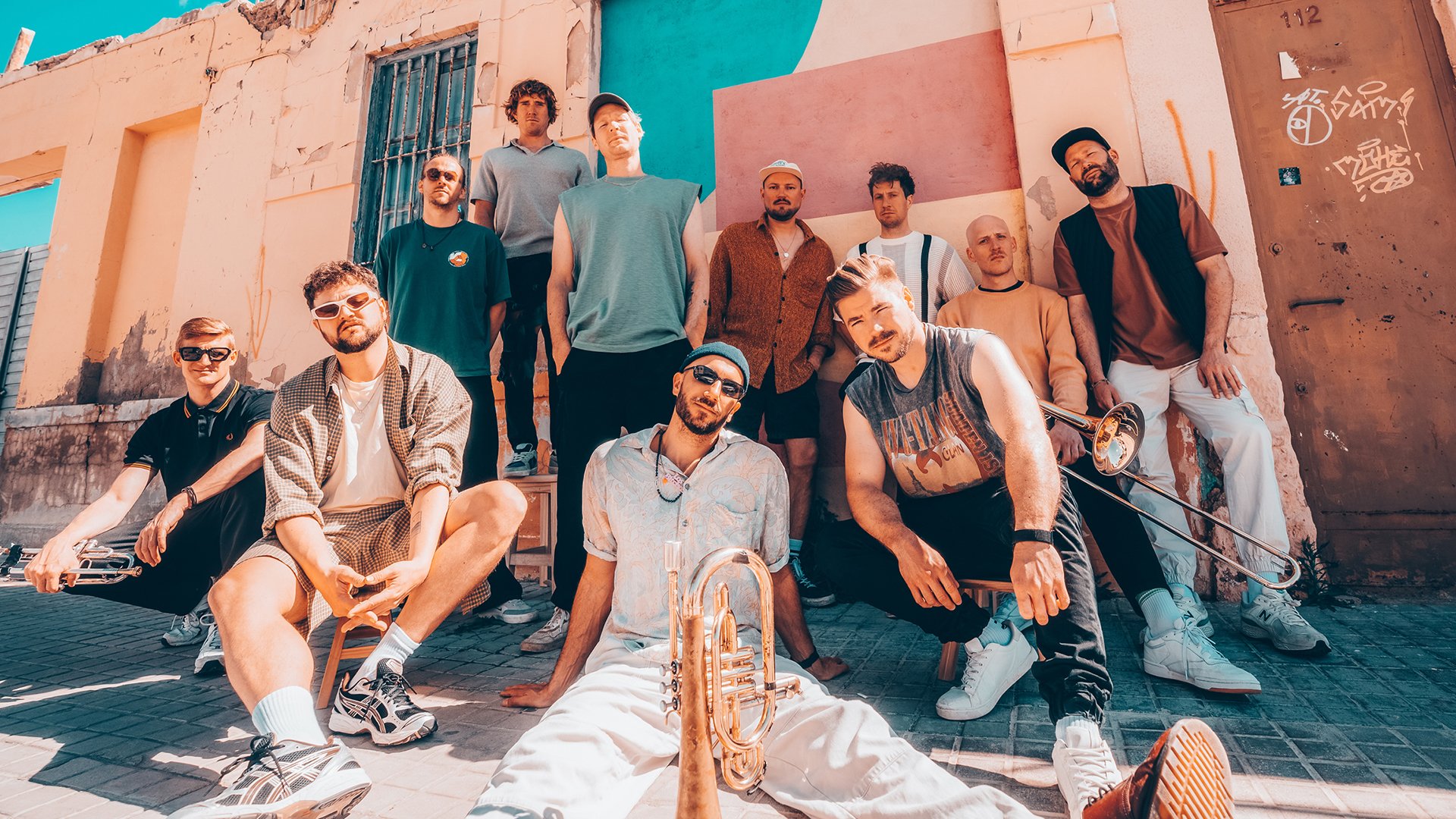 Bandfoto von Querbeat: Mehrere Personen sitzen und stehen im Freien vor einer farbigen Hausfassade mit Türen und Fenstern. Einige halten Blechblasinstrumente wie Trompete und Posaune. Legere Kleidung, warme Tageslichtstimmung, urbanes Umfeld. Promomotiv zur Live‑Show von Querbeat beim Tollwood Sommerfestival in München.