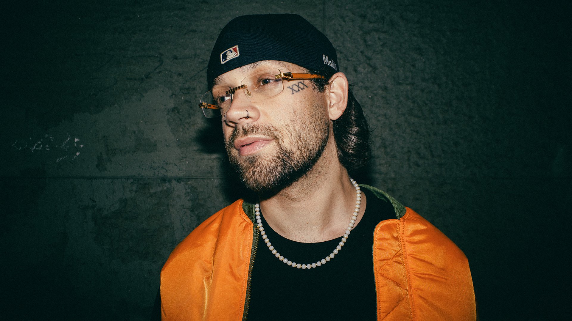 Foto für die Veranstaltung ‚FAYAN – VERLIEBT IN MICH TOUR 2026‘ mit einem Foto des Künstlers in einer orangefarbenen Jacke, angelehnt an eine grüne Wand.