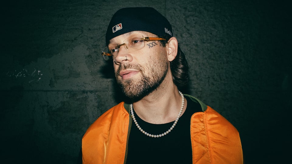 Foto für die Veranstaltung ‚FAYAN – VERLIEBT IN MICH TOUR 2026‘ mit einem Foto des Künstlers in einer orangefarbenen Jacke, angelehnt an eine grüne Wand.