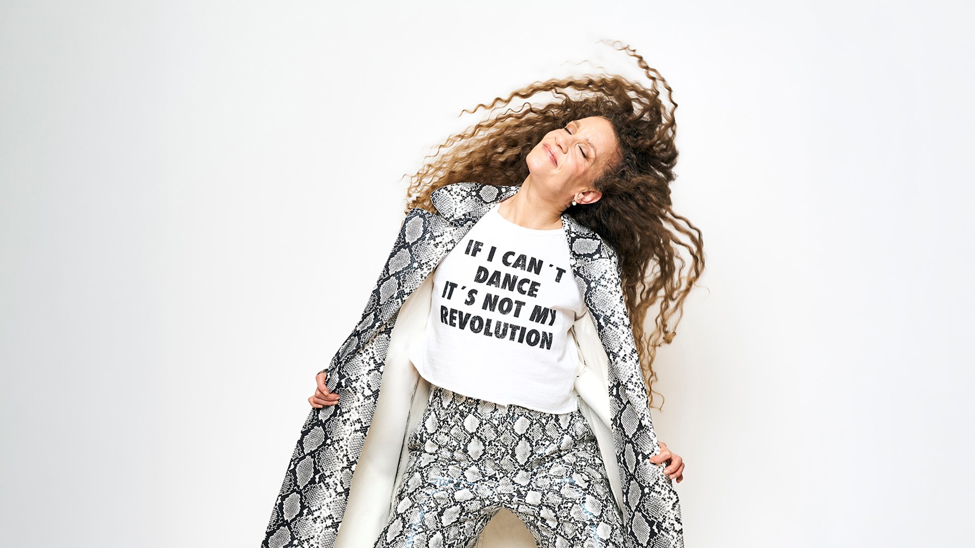 Amanda Marshall lächelnd in einer Tanzbewegung mit fliegendem Haar in schwarz-weiß gemusterter Hose und Schal sowie weißes Shirt mit Aufdruck If I cam´t dance it´s not my revolution, vor weißem Hintergrund