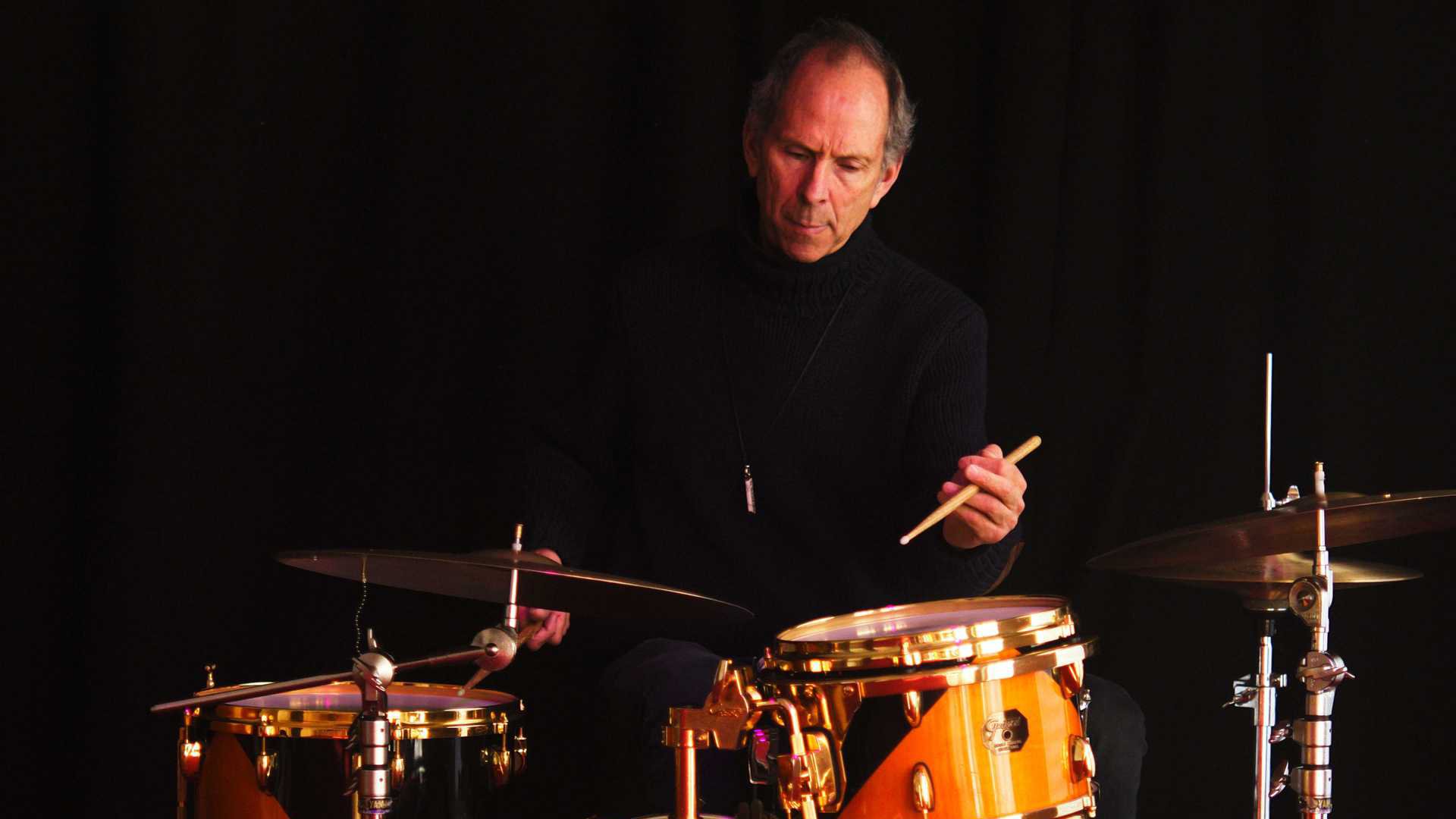 Rick Hollander in schwarzem Rollkragenpullover vor schwarzem Hintergrund, sitzend an einem Drumset mit Trommeln und Becken