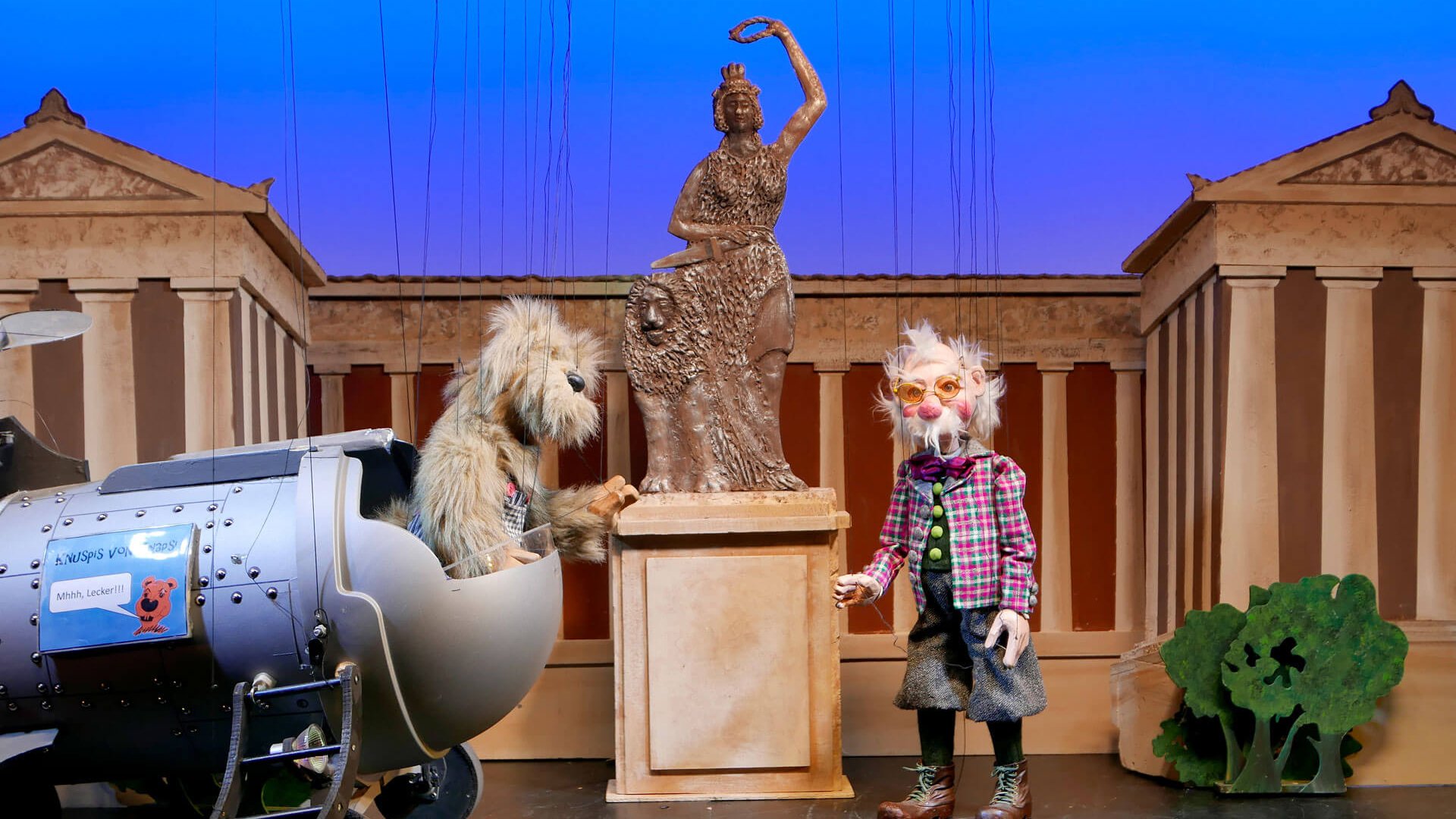 Puppenszene mit einem wuscheligen Hund in einer silbernen Raketenattrappe und einer älteren Marionettenfigur in karierter Kleidung; zwischen ihnen steht eine bronzefarbene Statue auf einem Sockel vor einem Bühnenbild mit klassizistischen Säulen und blauem Hintergrund