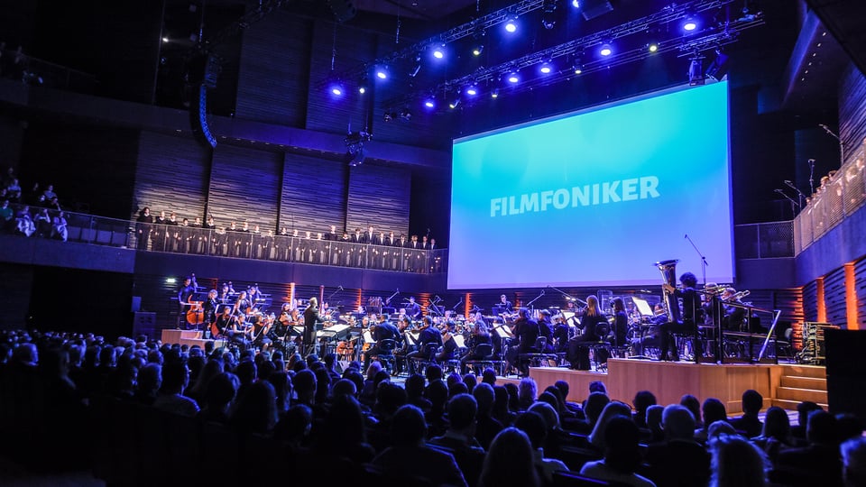 Bühnenfoto der Filmfoniker mit großem Symphonieorchester und Chor vor Publikum in einem Konzertsaal, darüber eine Leinwand mit dem Schriftzug Filmfoniker in blauem Licht