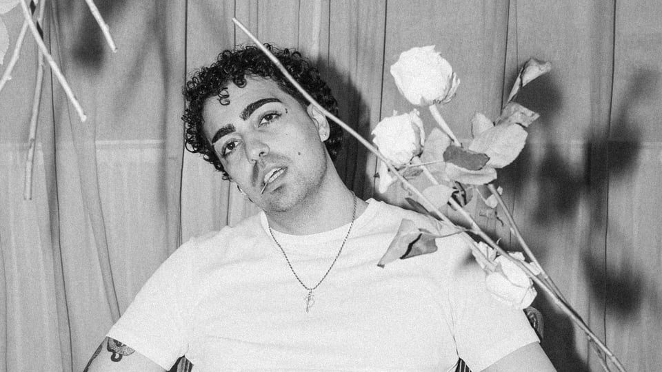 Schwarz‑weißes Porträt einer Person mit kurzen lockigen Haaren und hellem T‑Shirt vor Vorhang, hält einen Blumenstrauß mit Rosen
