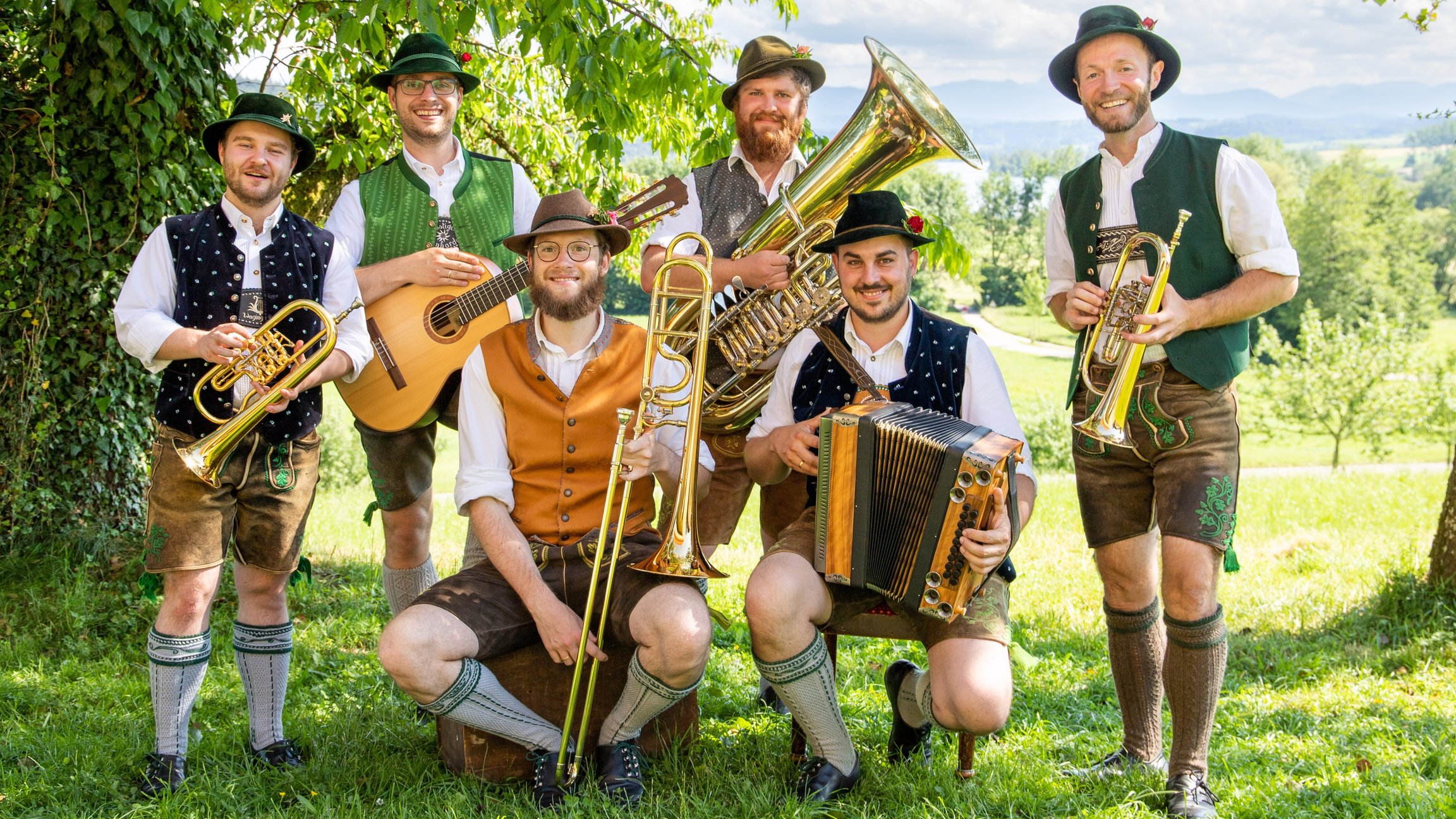 Sechs Musiker mit traditionellen Instrumenten stehen auf einer grünen Wiese mit Bäumen im Hintergrund