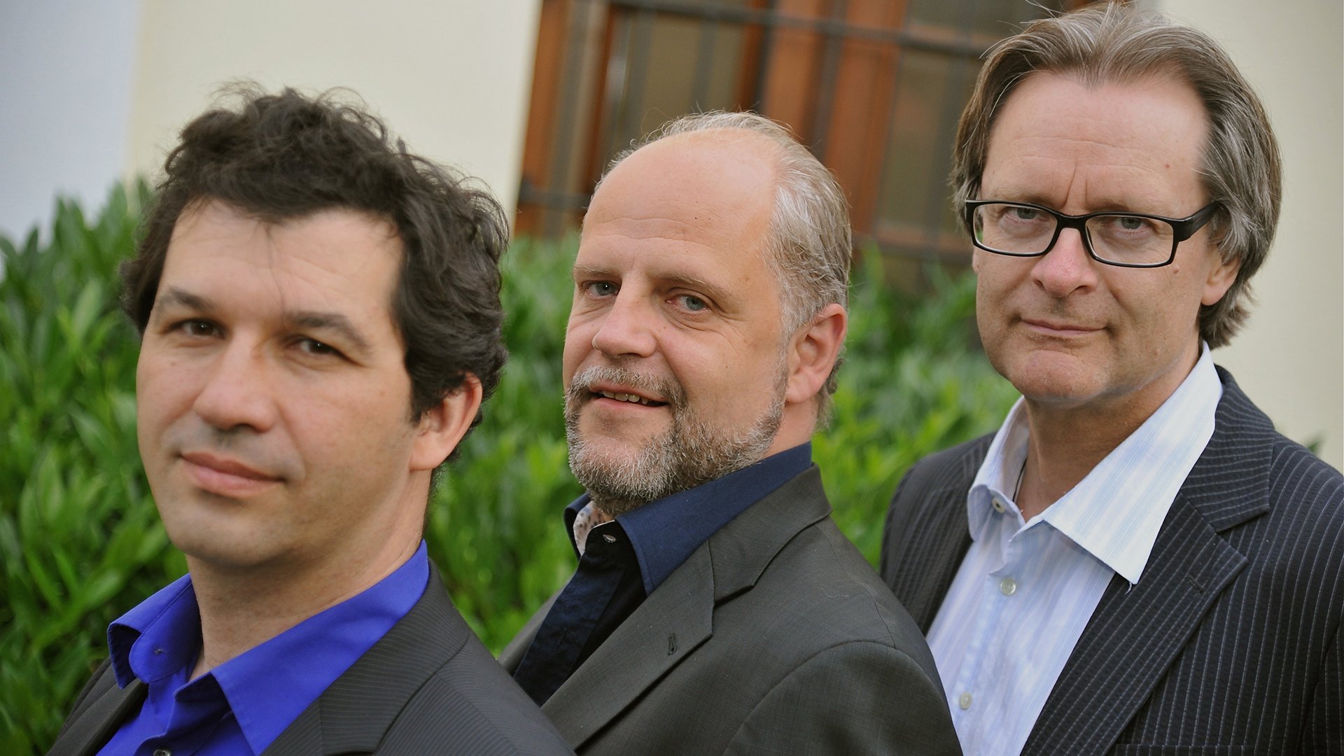 Porträt von drei Musikern in eleganter Kleidung vor grünem Hintergrund im Freien.