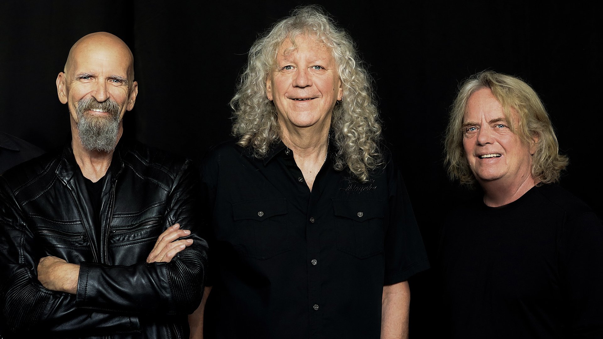 Die drei Mitglieder der Band Saga stehe in dunkler Kleidung vor einem dunklen Hintergrund.