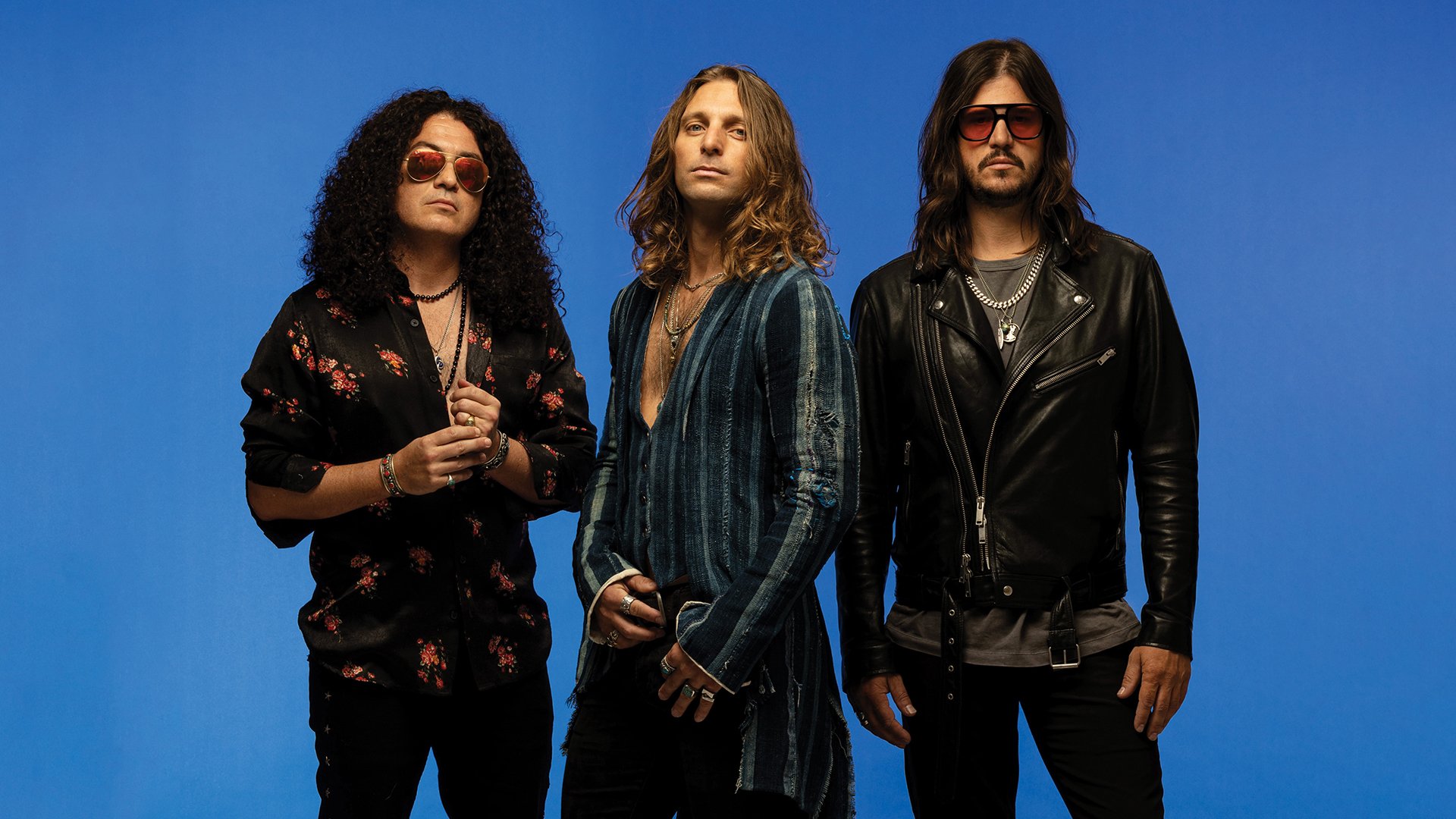 Bandfoto von Dirty Honey mit drei Bandmitgliedern vor blauem Studiohintergrund, in rocktypischer Kleidung mit langen Haaren, Sonnenbrillen, Lederjacke und gemusterten Hemden