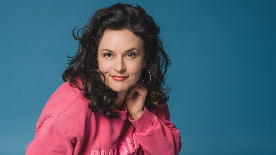 Angela Ascher in pinkem Sweatshirt lächelt in die Kamera, vor blauem Hintergrund