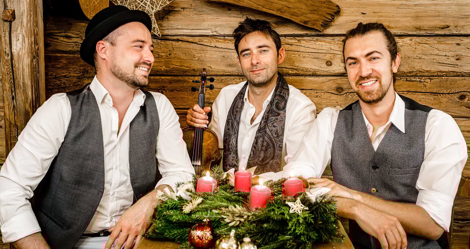 Drei Männer vor Holzwand mit Adventskranz und brennenden Kerzen