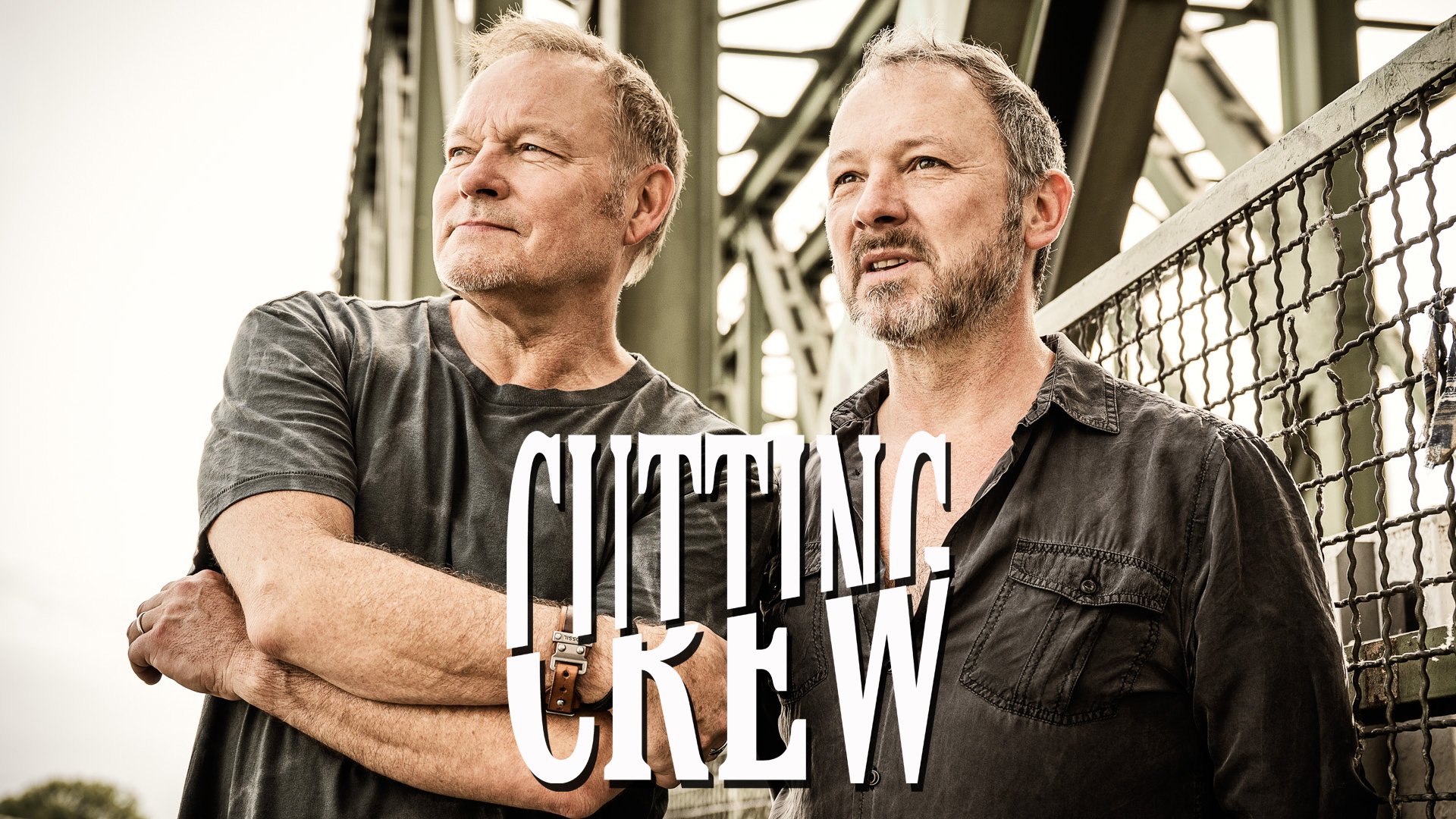Eventmotiv der Band Cutting Crew: Zwei Personen stehen nebeneinander im Außenbereich vor einer Metall‑Brückenkonstruktion mit Geländer. Beide tragen dunkle, legere Kleidung und blicken seitlich aus dem Bild. Zentrisch im Vordergrund ist der Schriftzug „CUTTING CREW“ zu sehen. Natürliches Tageslicht, urban‑industrielle Umgebung; Promomotiv für ein Live‑Konzert.
