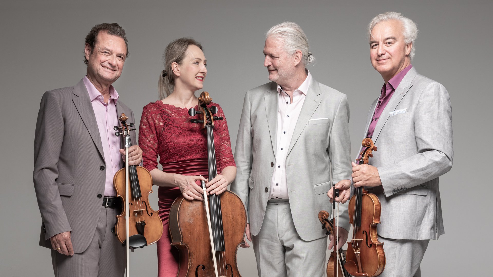 Das Modern String Quartetts mit vier Personen in eleganter Konzertkleidung vor hellgrauem Hintergrund, drei mit Violine und Violoncello in den Händen, eine Person im roten Spitzenkleid zwischen drei Personen in hellen Anzügen.