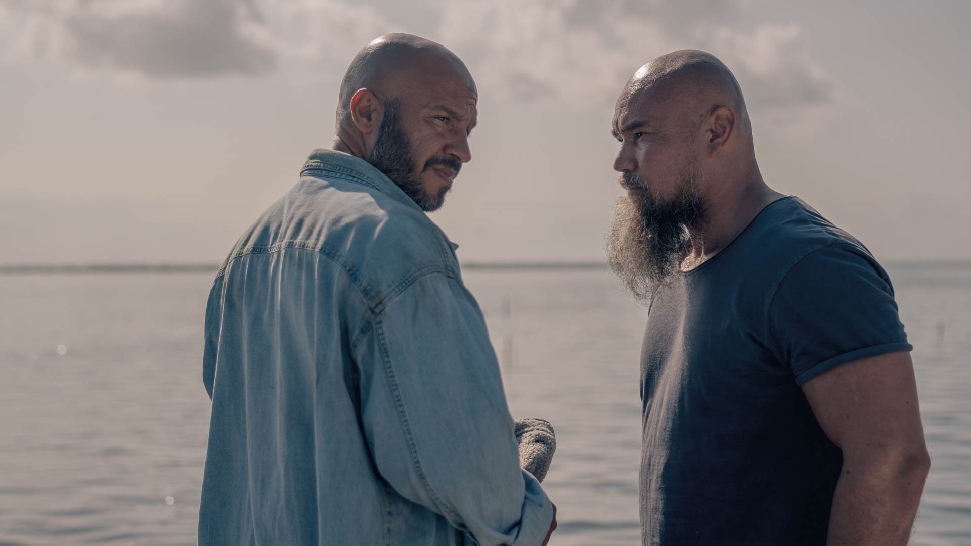 Zwei Männer mit Glatze und Bart unterhalten sich. Im Hintergrund ist das Meer zu sehen.