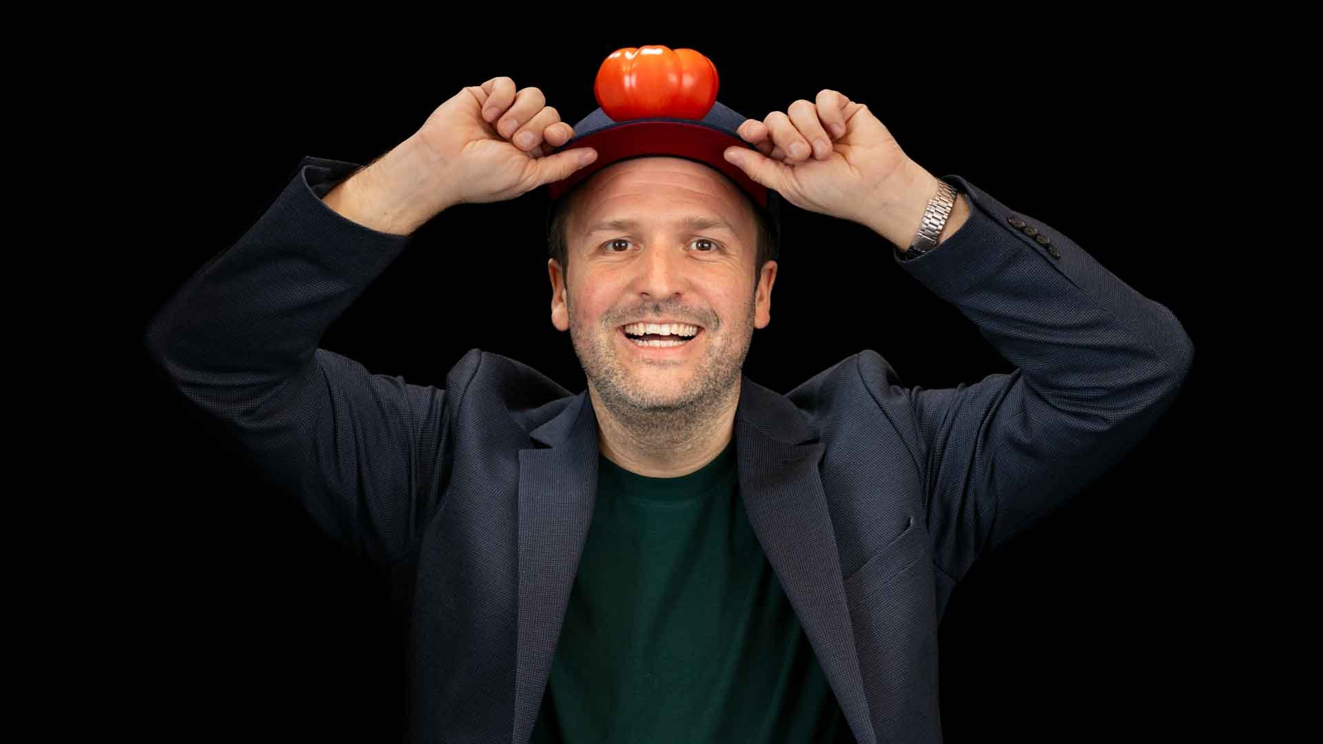 Porträt einer Person mit dunklem Sakko und Baseballkappe vor schwarzem Hintergrund. Auf der Kappe sitzt ein kleiner orangefarbener Kürbis, während die Person den Schirm der Kappe mit beiden Händen festhält.