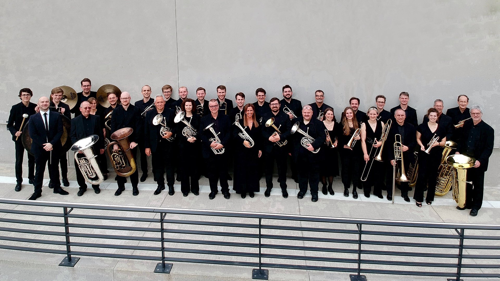 Im Vordergrund verläuft ein Metallgeländer entlang einer Stufe, dahinter steht die Brass Band München in schwarzer Kleidung mit Blechblasinstrumenten vor einer hellen Wand