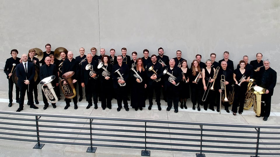 Im Vordergrund verläuft ein Metallgeländer entlang einer Stufe, dahinter steht die Brass Band München in schwarzer Kleidung mit Blechblasinstrumenten vor einer hellen Wand