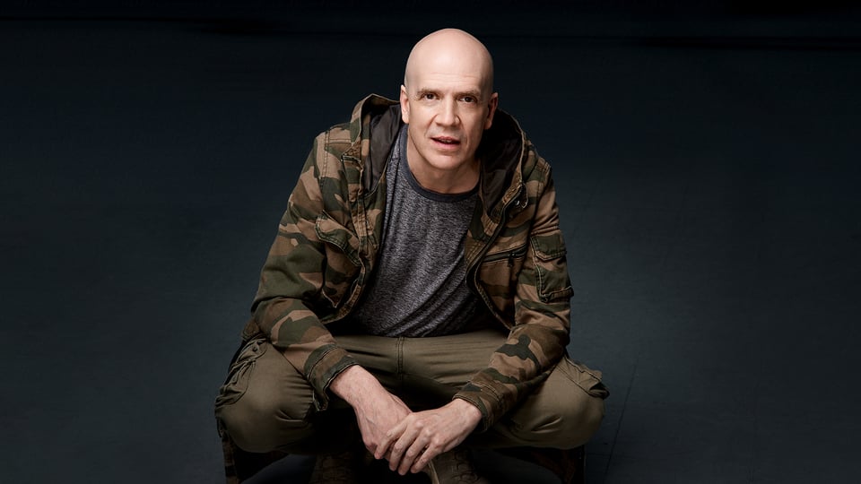 Devin Townsend in Tarnjacke sitzt nach vorne gebeugt vor dunklem Hintergrund