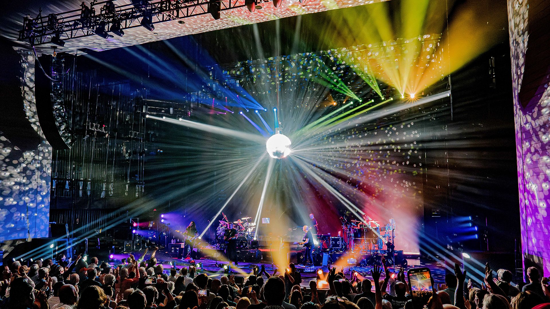 Bühnenfoto von Brit Floyd auf einer Konzertbühne vor Publikum, mit großer Discokugel über der Bühne, farbigen Lichtstrahlen und Projektionen sowie mehreren Musikern unter spektakulärer Lichtshow