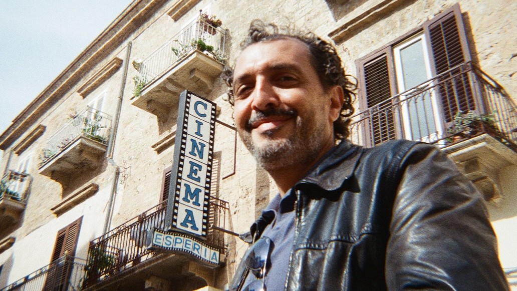 Der Künstler Kid Francescoli steht vor einer altertümlichen Hauswand mit Balkonen sowie einem Schild mit der Aufschrift Cinema