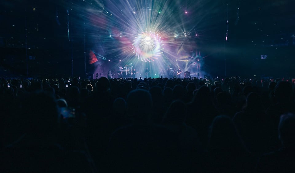 Ein Konzert mit strahlendem Licht, welches von der Bühne ausgeht, das Publikum ist von hinten zu sehen, wie es auf die Bühne schaut.