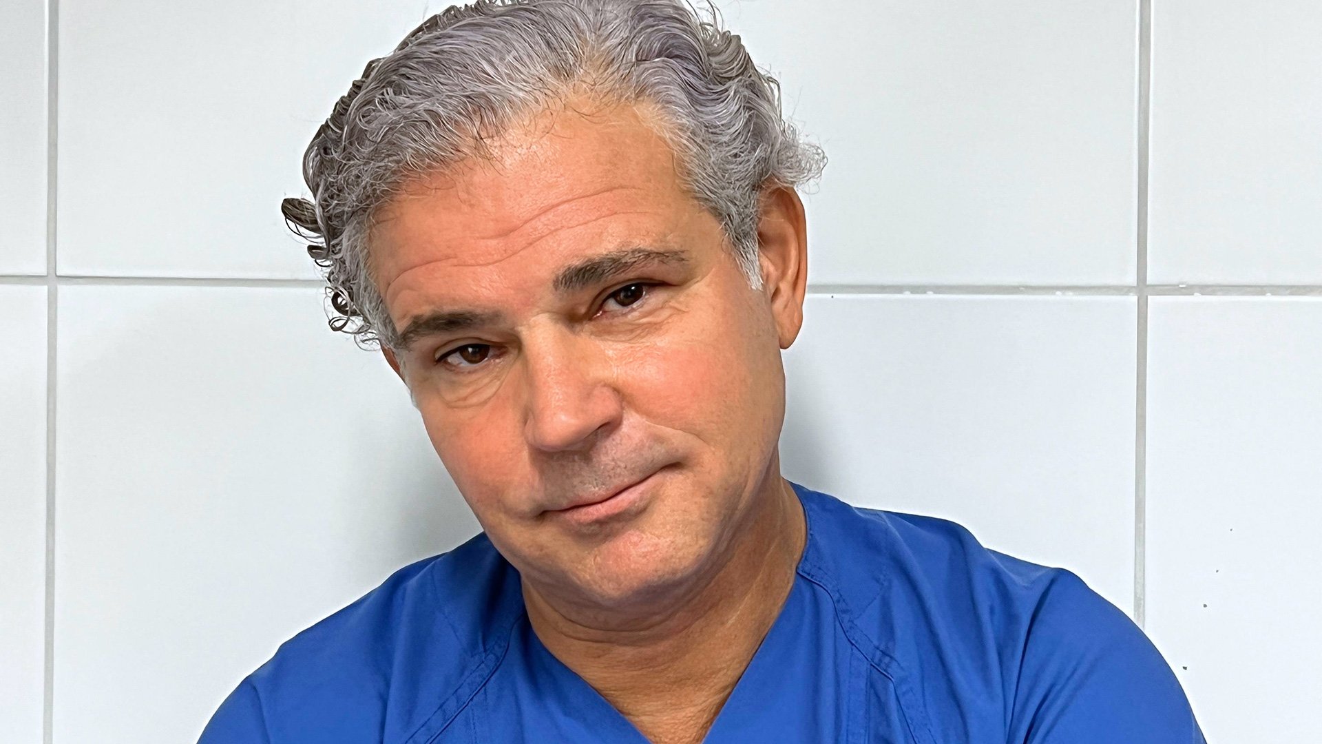 Foto von Michael Tsokos mit blauem Krankenhaushemd vor einer weißen Kachelwand wie im Krankenhaus