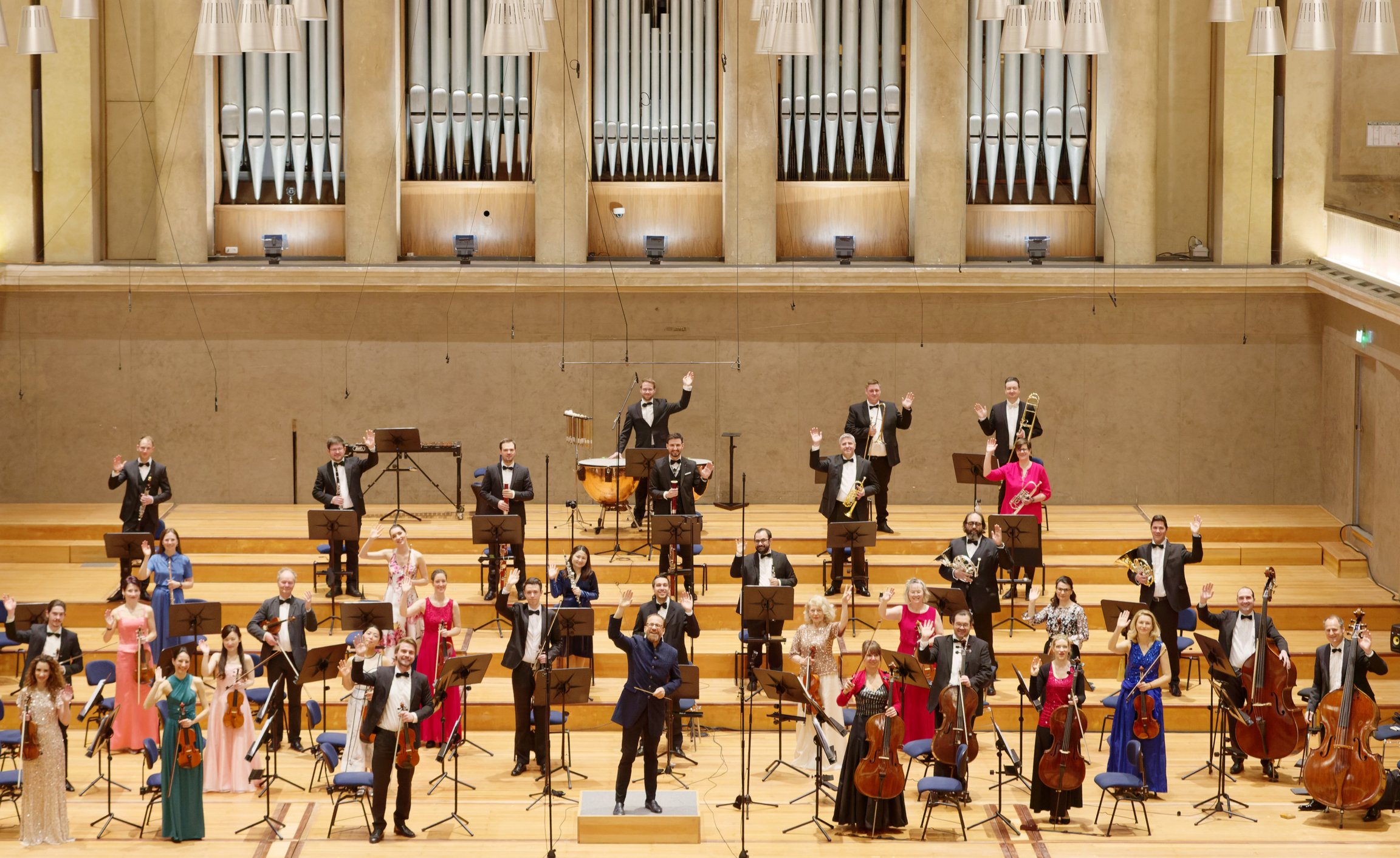 Großes Orchester auf einer Konzertbühne, Dirigent vorne mittig, Musiker mit Streich- und Blasinstrumenten, mit großer Orgel im Hintergrund.