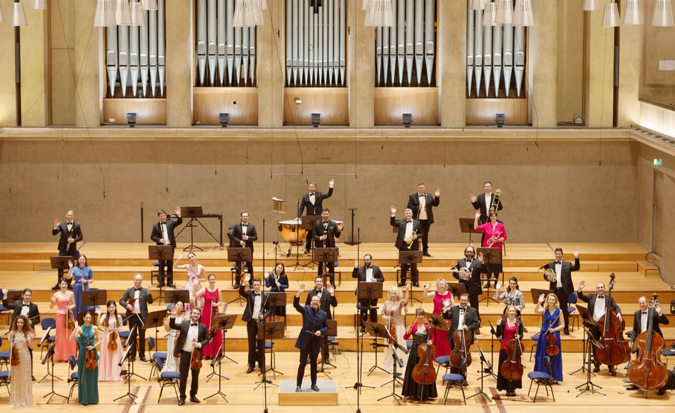 Großes Orchester auf einer Konzertbühne, Dirigent vorne mittig, Musiker mit Streich- und Blasinstrumenten, mit großer Orgel im Hintergrund.