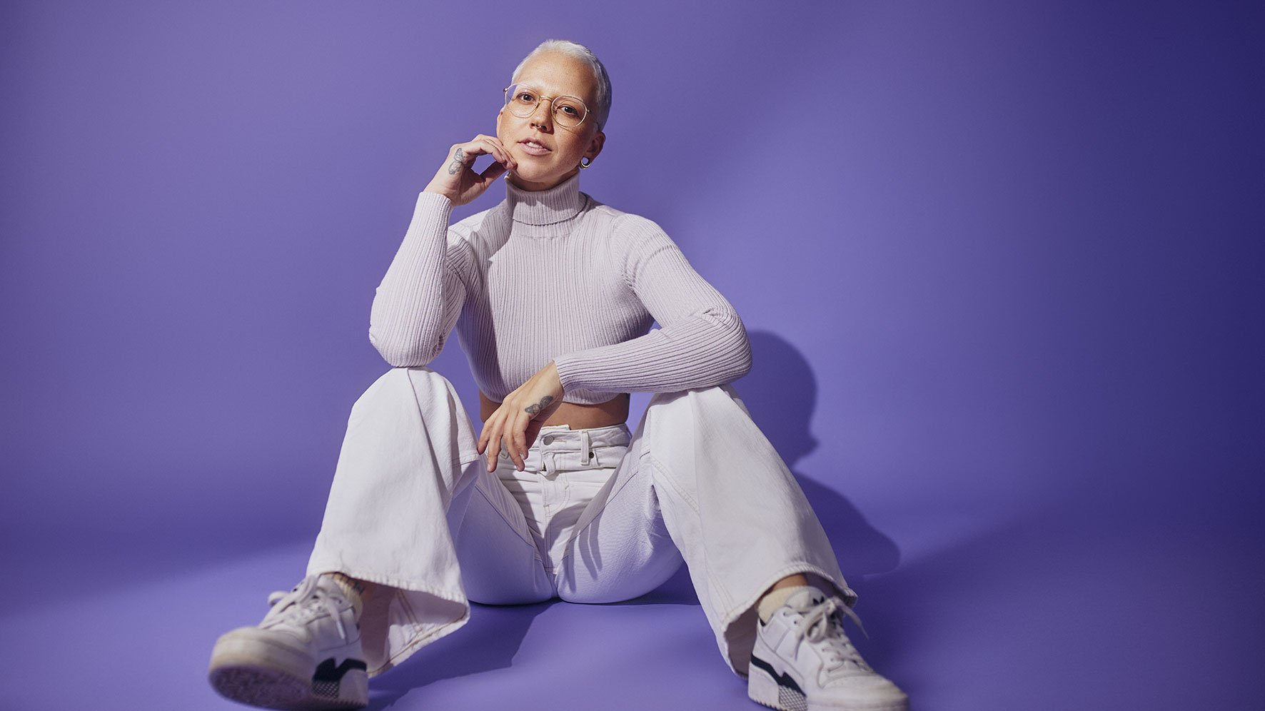 Stefanie Heinzmann mit sehr kurzen hellen Haaren und runder Brille sitzt vor violettem Hintergrund. Heller Rollkragenpullover, weiße Hose und weiße Turnschuhe prägen die Porträtaufnahme.