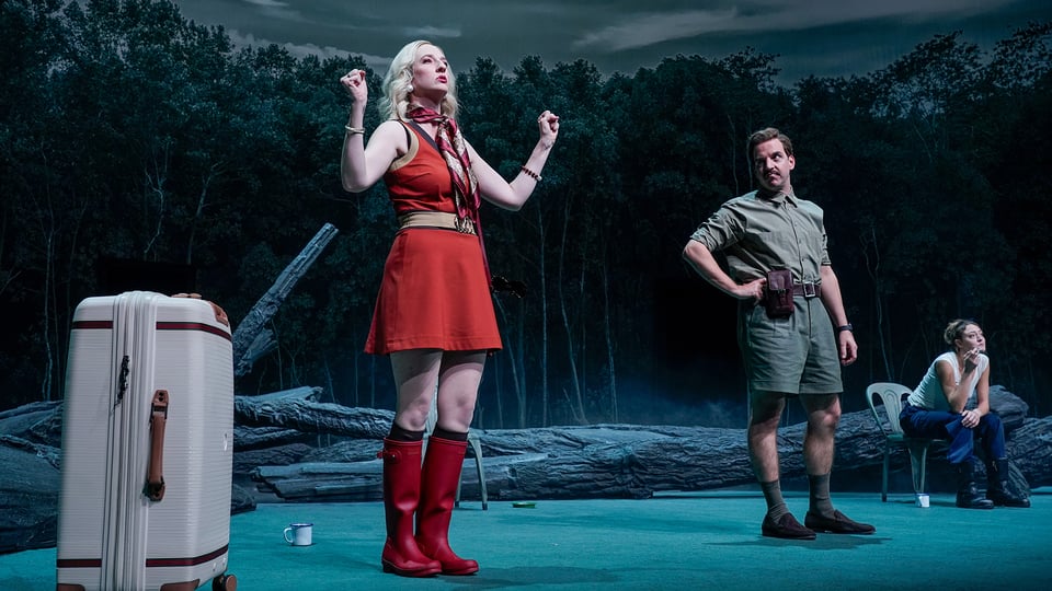 Eine rot gekleidete Dame und ein Herr Stehen auf einer Bühne mit einem Wald im Hintergrund, ein Koffer vorne